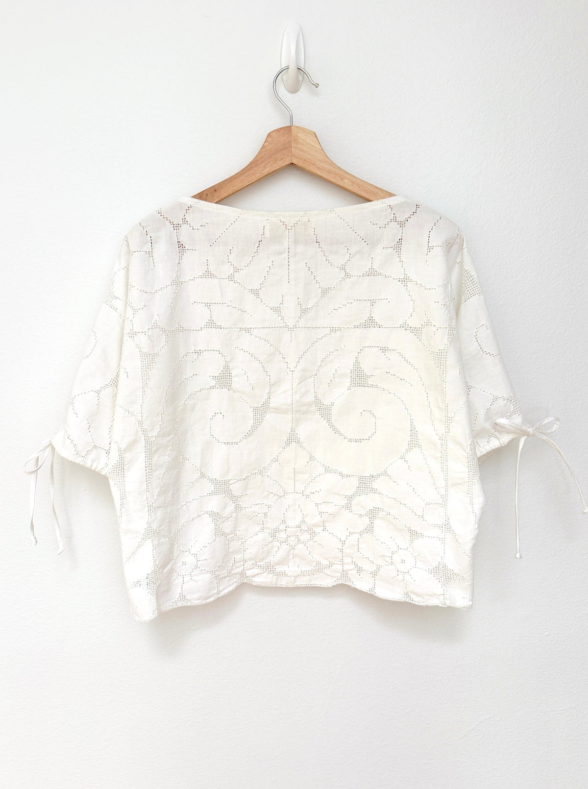 A white lace short-sleeve blouse with a floral damask pattern and tie details on the sleeves, hanging on a wooden hanger against a plain white wall.