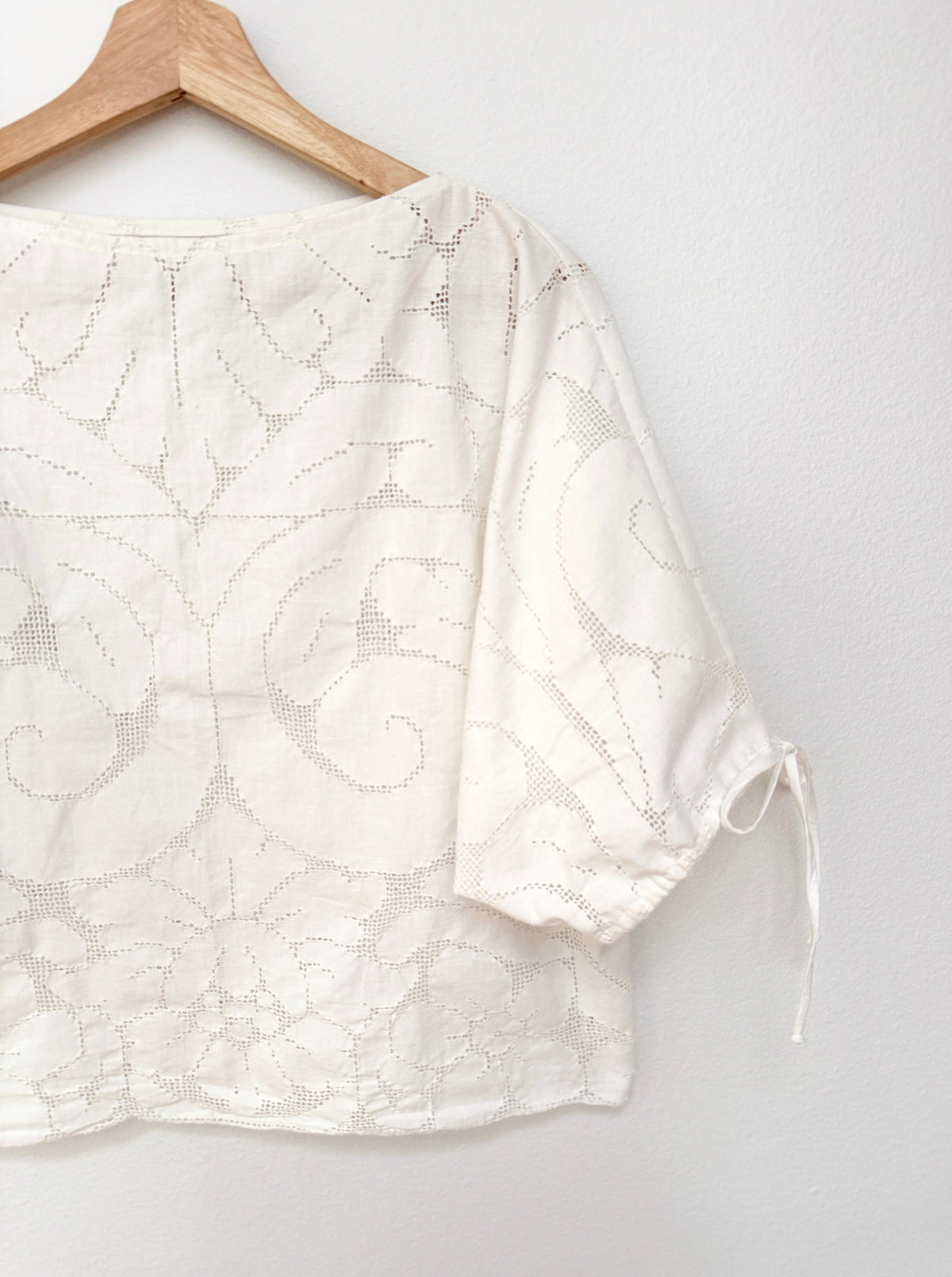 A white embroidered blouse with short puff sleeves and side ties, hanging on a wooden hanger against a white wall.