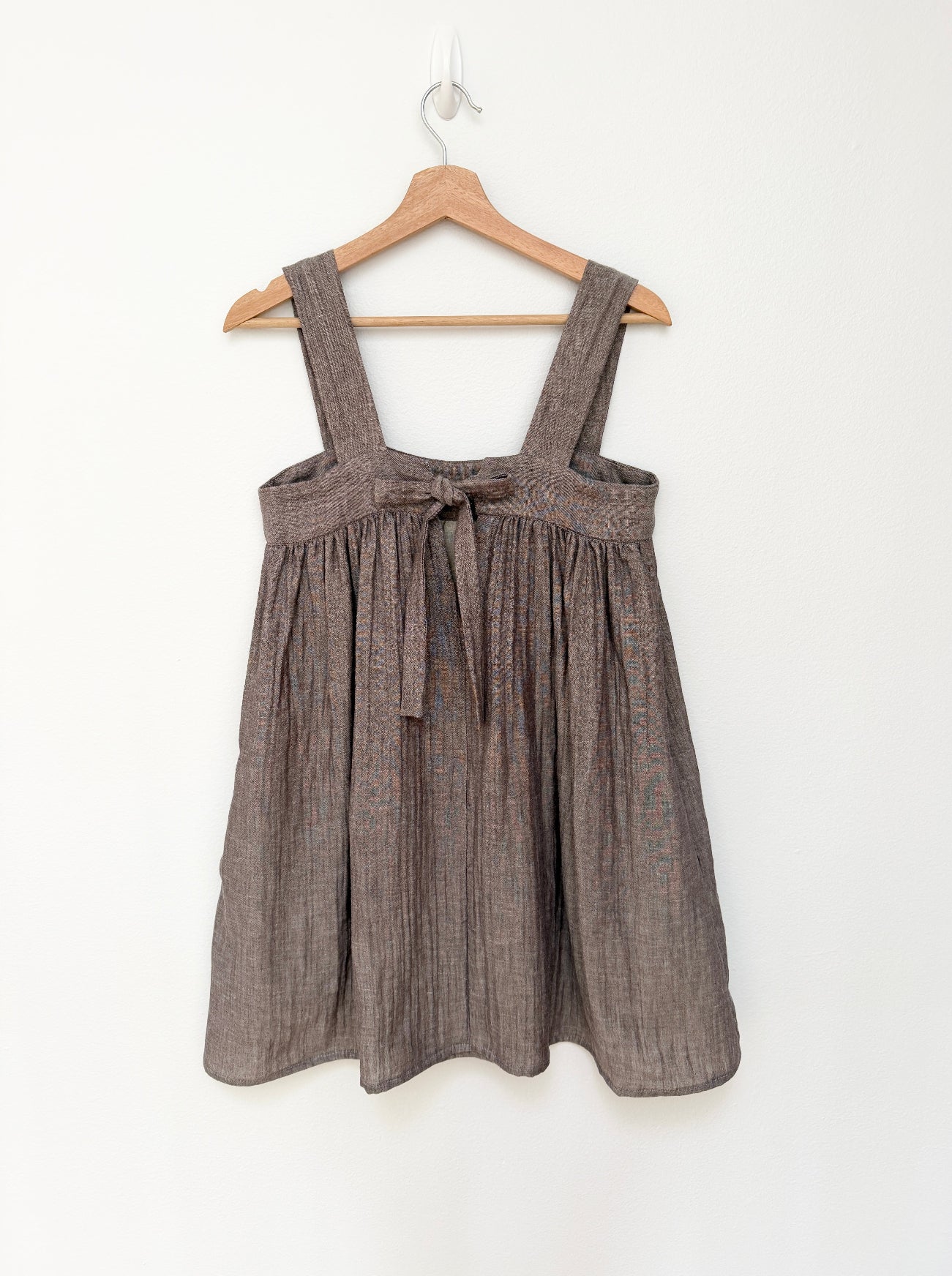 A sleeveless brown cotton dress with wide shoulder straps and a front tie detail, hanging on a wooden hanger against a plain white wall.