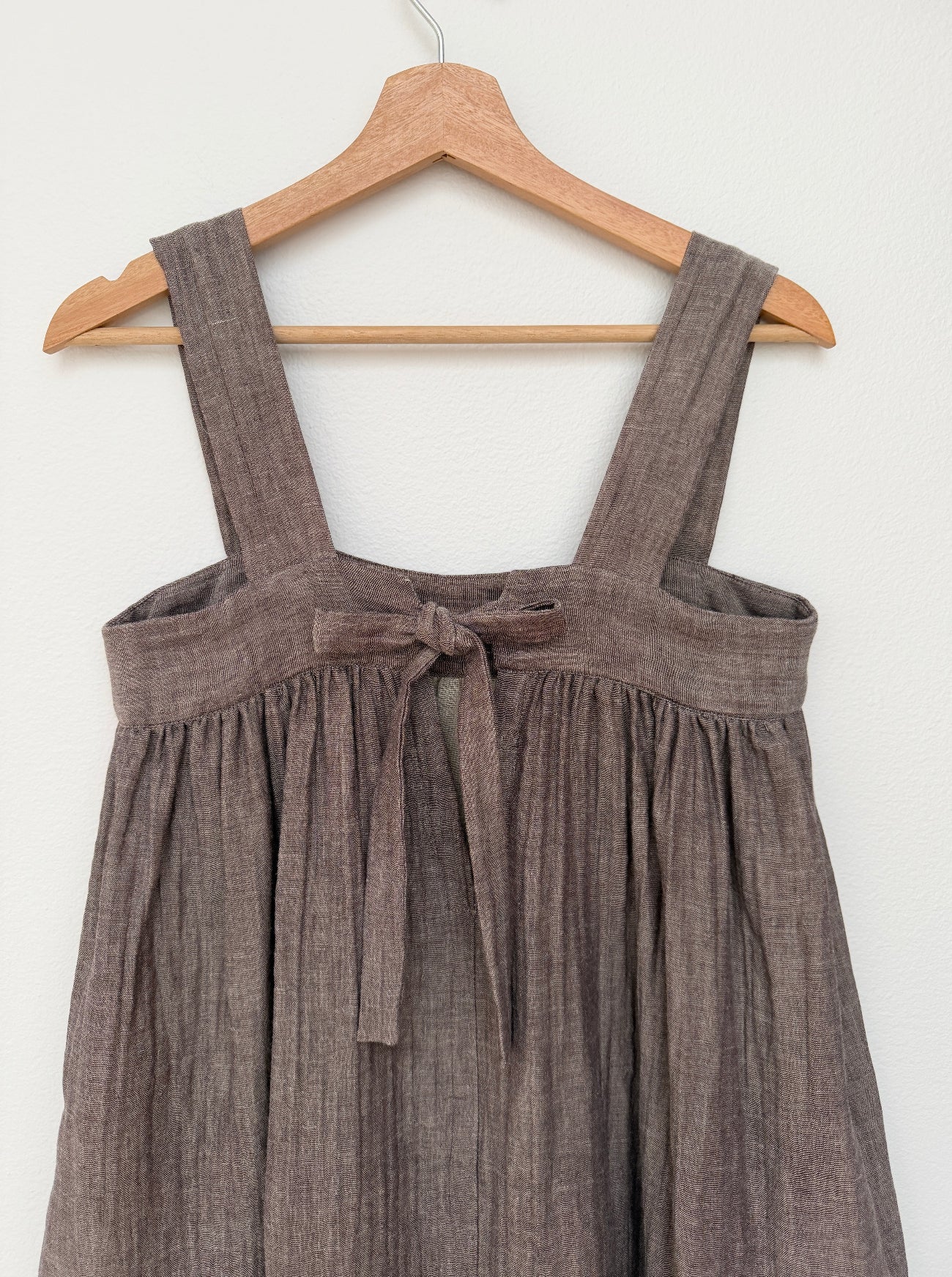 A sleeveless brown cotton dress with wide shoulder straps and a front tie detail, hanging on a wooden hanger against a plain white wall.