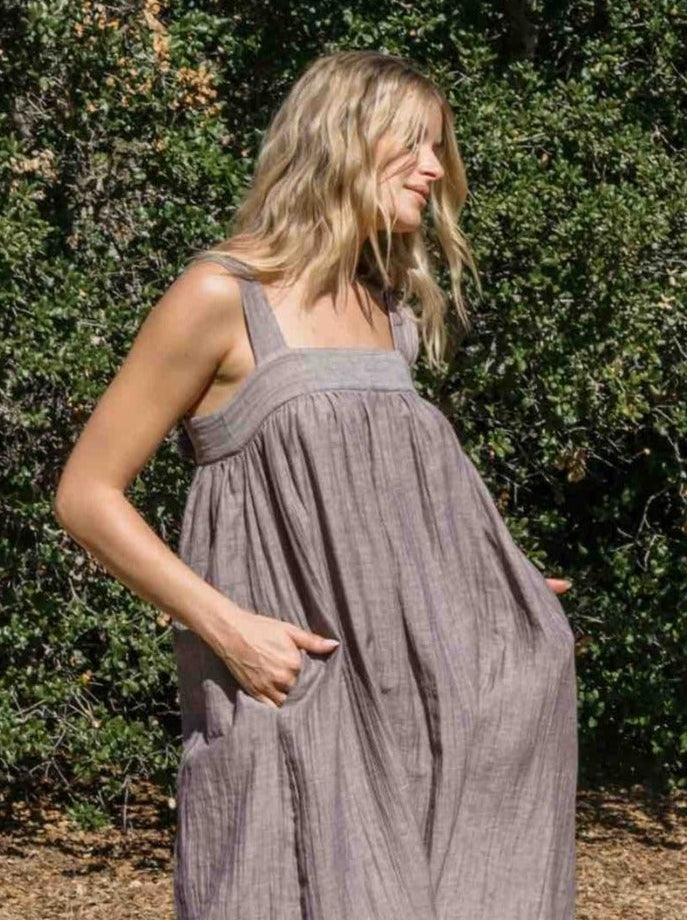 A woman wearing a loose, sleeveless brown midi dress with pockets, standing outdoors on a dirt path with green bushes in the background.