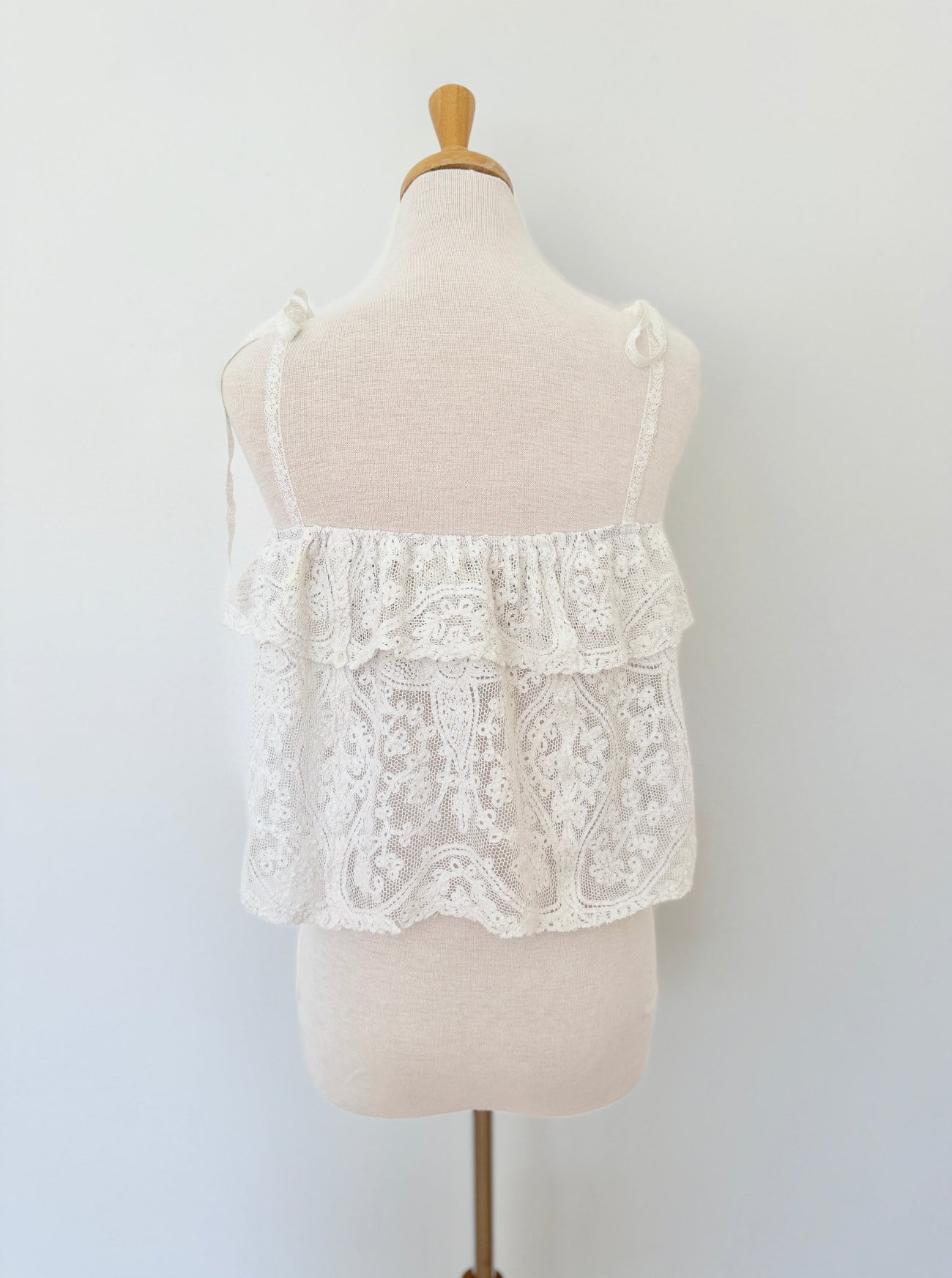 An off-white lace camisole top with ruffles and thin shoulder ties, displayed on a cream-colored fabric dress form mannequin against a plain white wall.