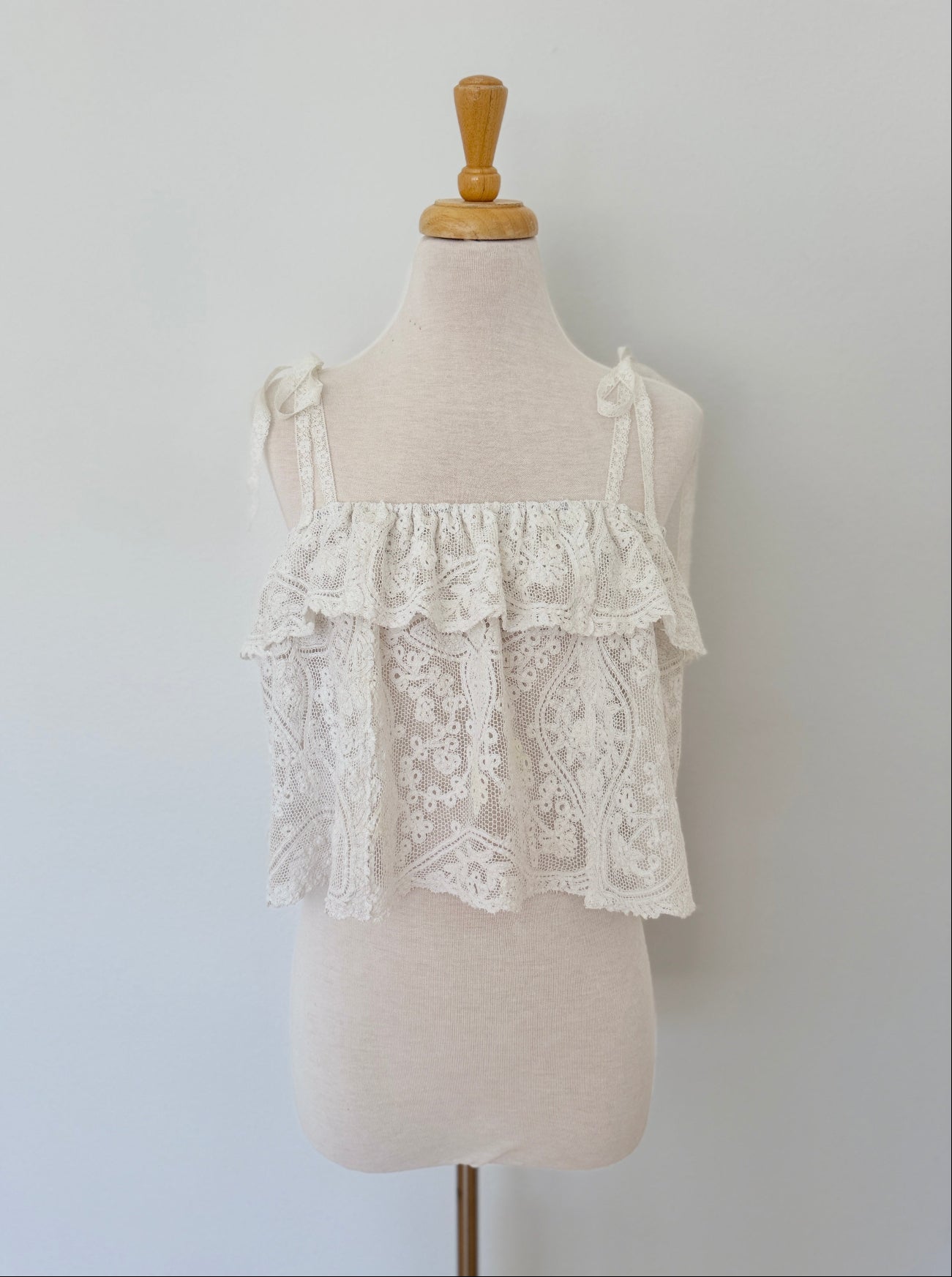 An off-white lace ruffled camisole top displayed on a cream fabric dress form mannequin against a light gray wall.