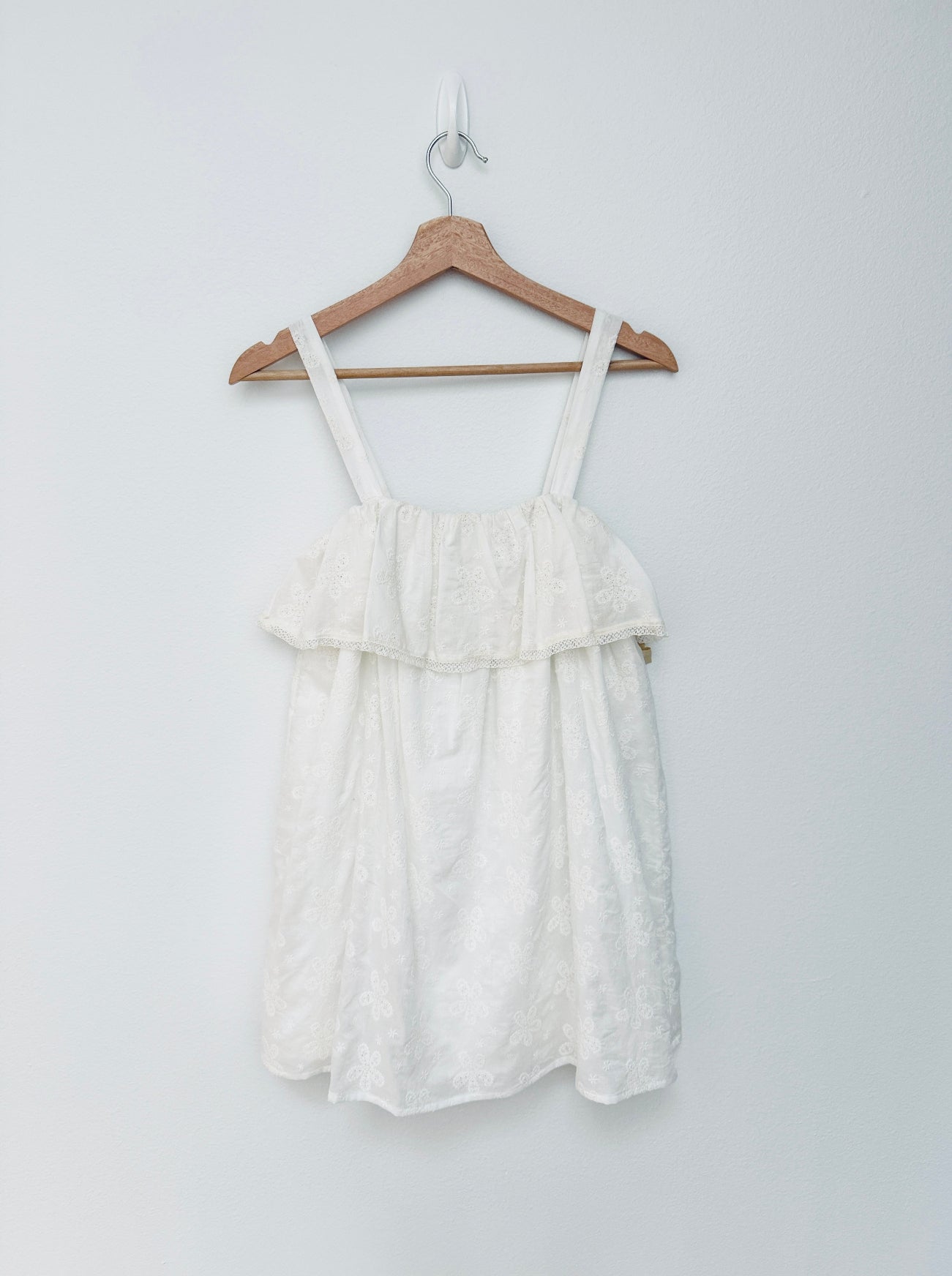 An white textured mini dress with thin shoulder straps and a ruffled tiered neckline, hanging on a wooden hanger against a plain light gray wall.
