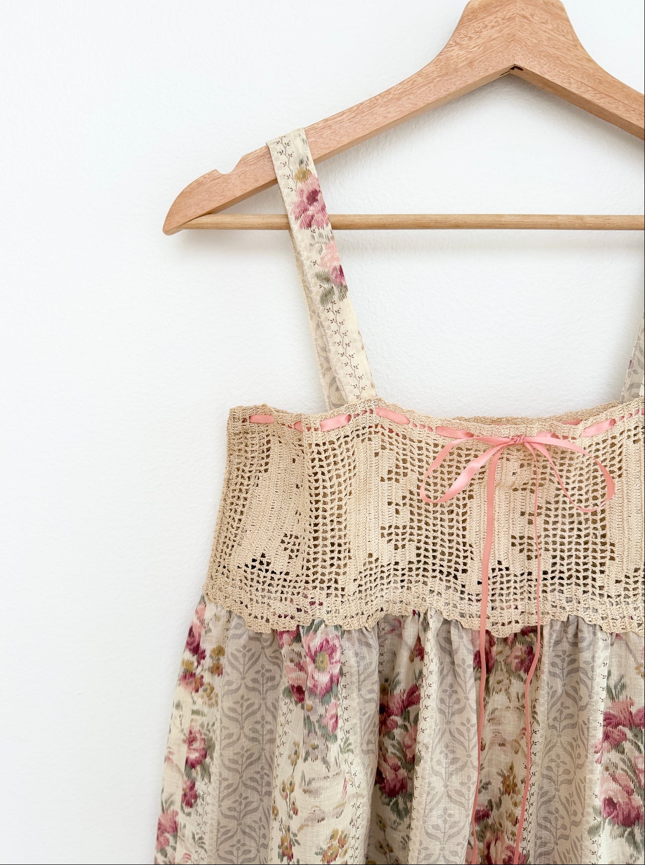 A close-up of a floral and lace dress hanging on a wooden hanger against a plain white wall. The dress features a cream base with pink and purple floral patterns, a beige crochet lace bodice, and thin shoulder straps.