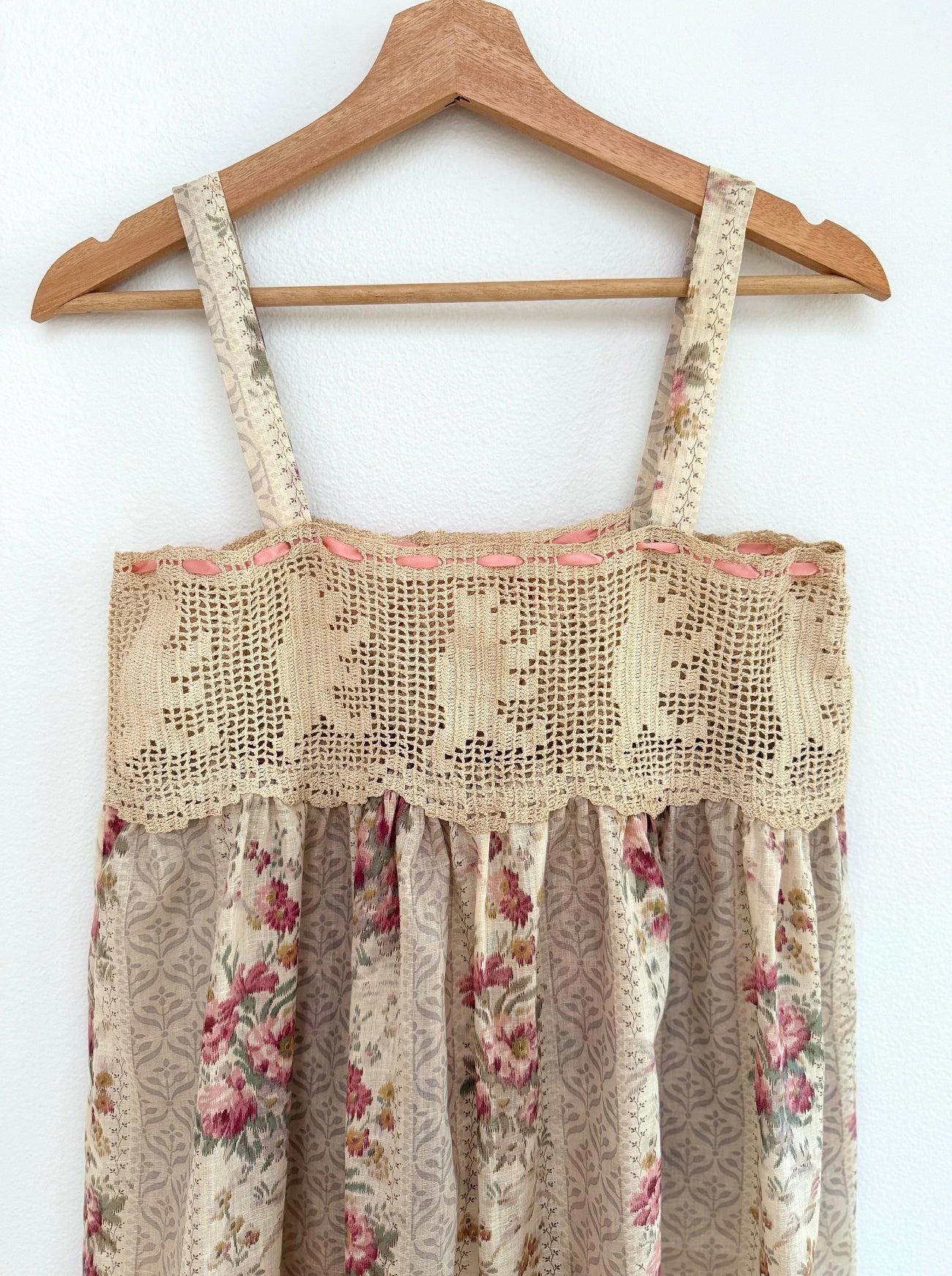 A sleeveless cream and beige floral dress with a crochet lace yoke and patterned skirt, hanging on a wooden hanger against a white wall.