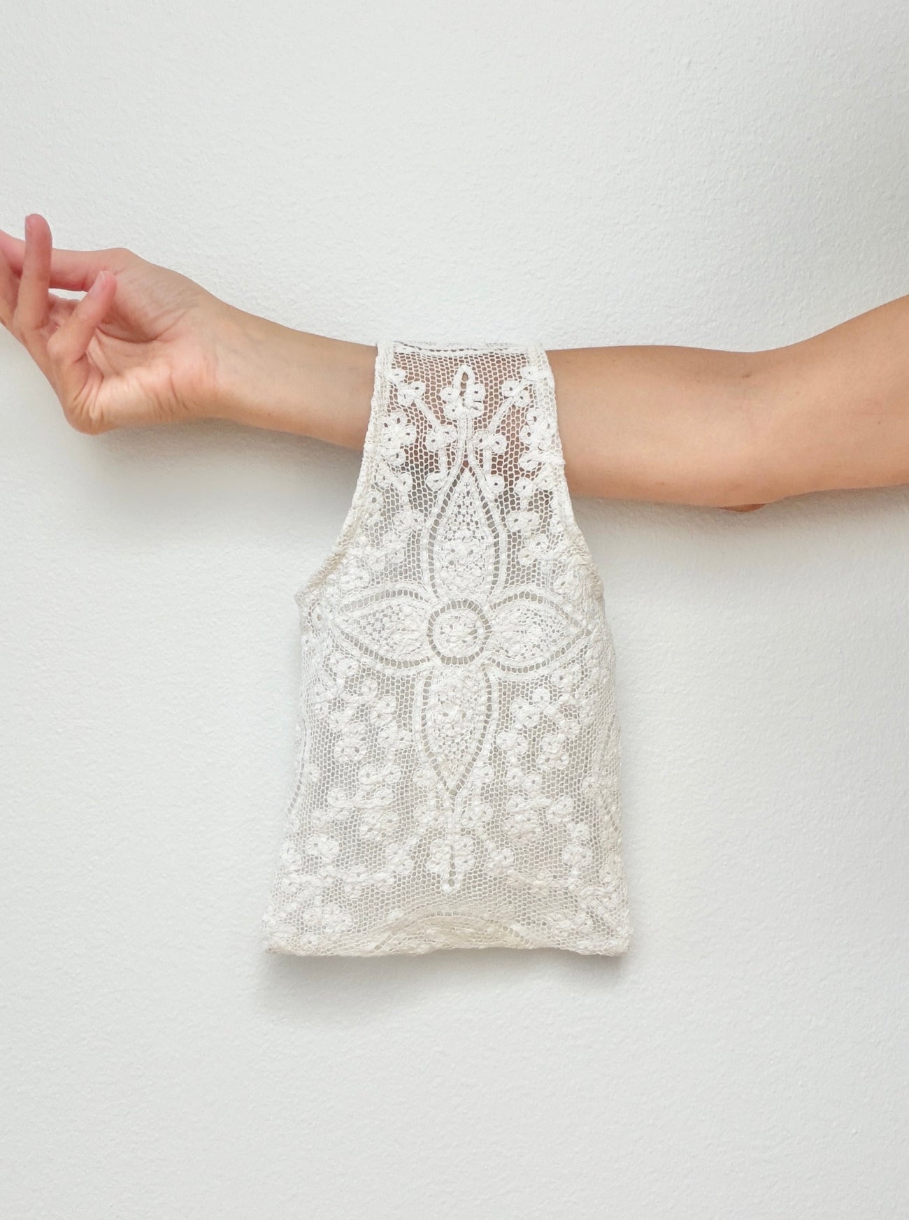 White lace sheer wristlet bag held up by a person's arm against a plain light gray wall.