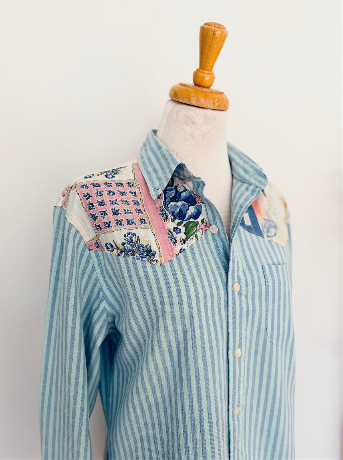 A light blue and white vertically striped button-down shirt with floral and patterned yoke panels on the shoulders, displayed on a cream dress form with a wooden neck cap against a plain white background.