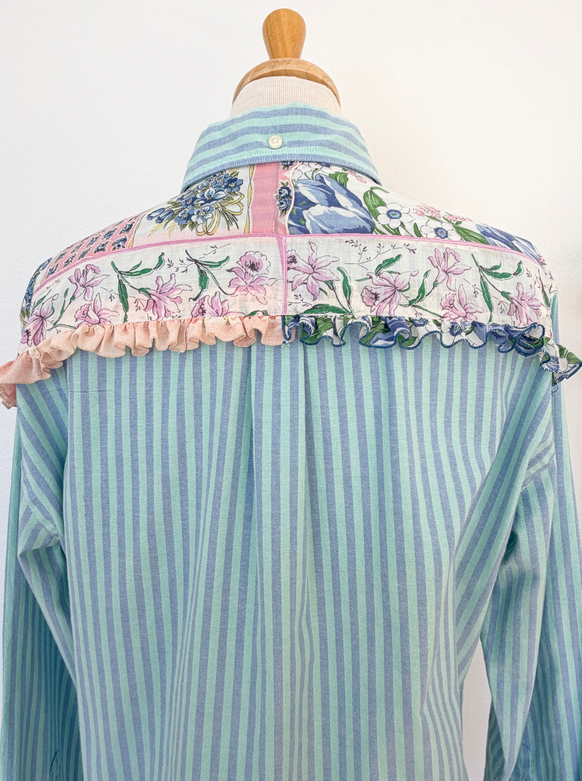 A light blue and white vertically striped button-down shirt with a decorative floral and ruffled yoke panel on the back, displayed on a dress form against a plain white background.