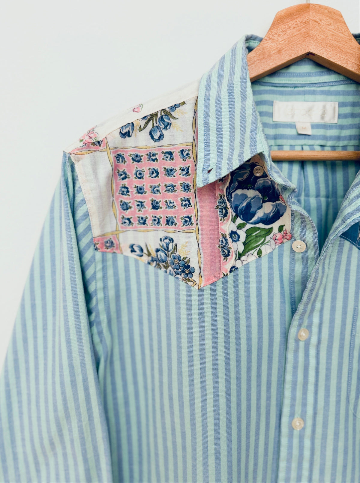 A light blue and white striped button-down shirt with a unique patchwork collar made from floral and patterned fabrics, hanging on a wooden hanger against a light gray background.