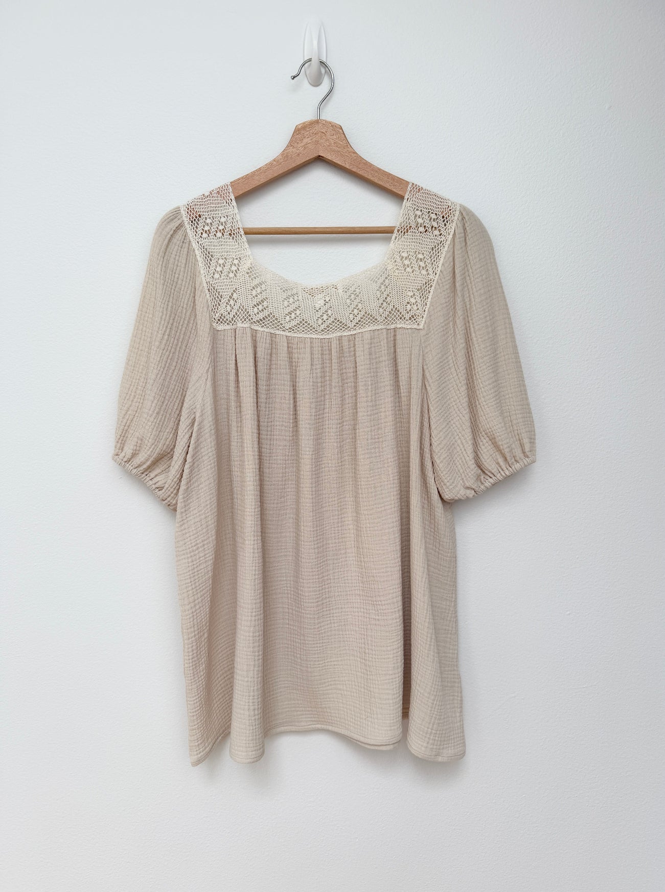 A beige short-sleeve mini dress with a textured waffle-like fabric, featuring a square neckline with white crochet lace trim and puff sleeves, hanging on a wooden hanger against a plain white wall.