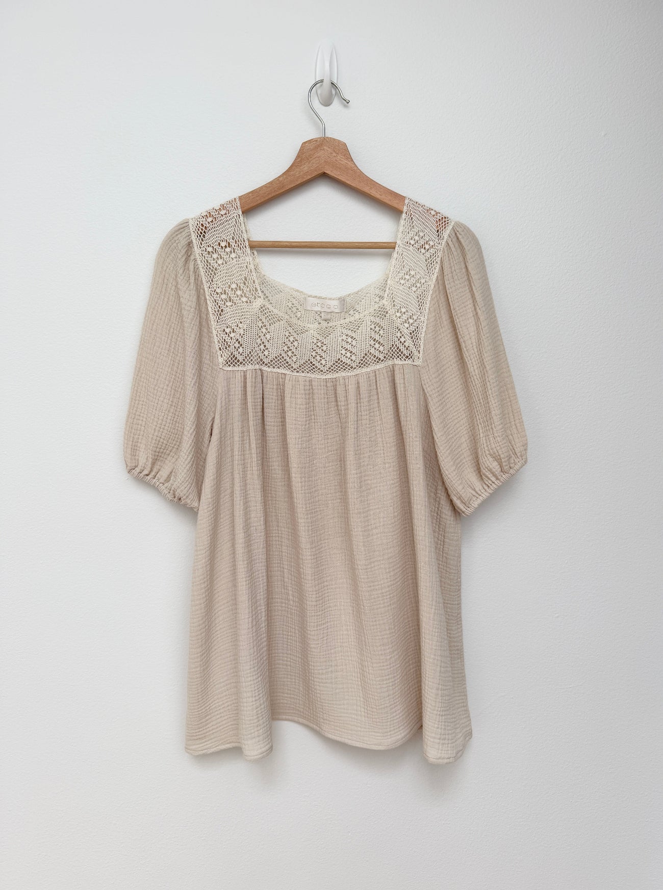 A beige short-sleeve mini dress with a textured waffle-weave body and a decorative lace yoke, hanging on a wooden hanger against a plain white wall.