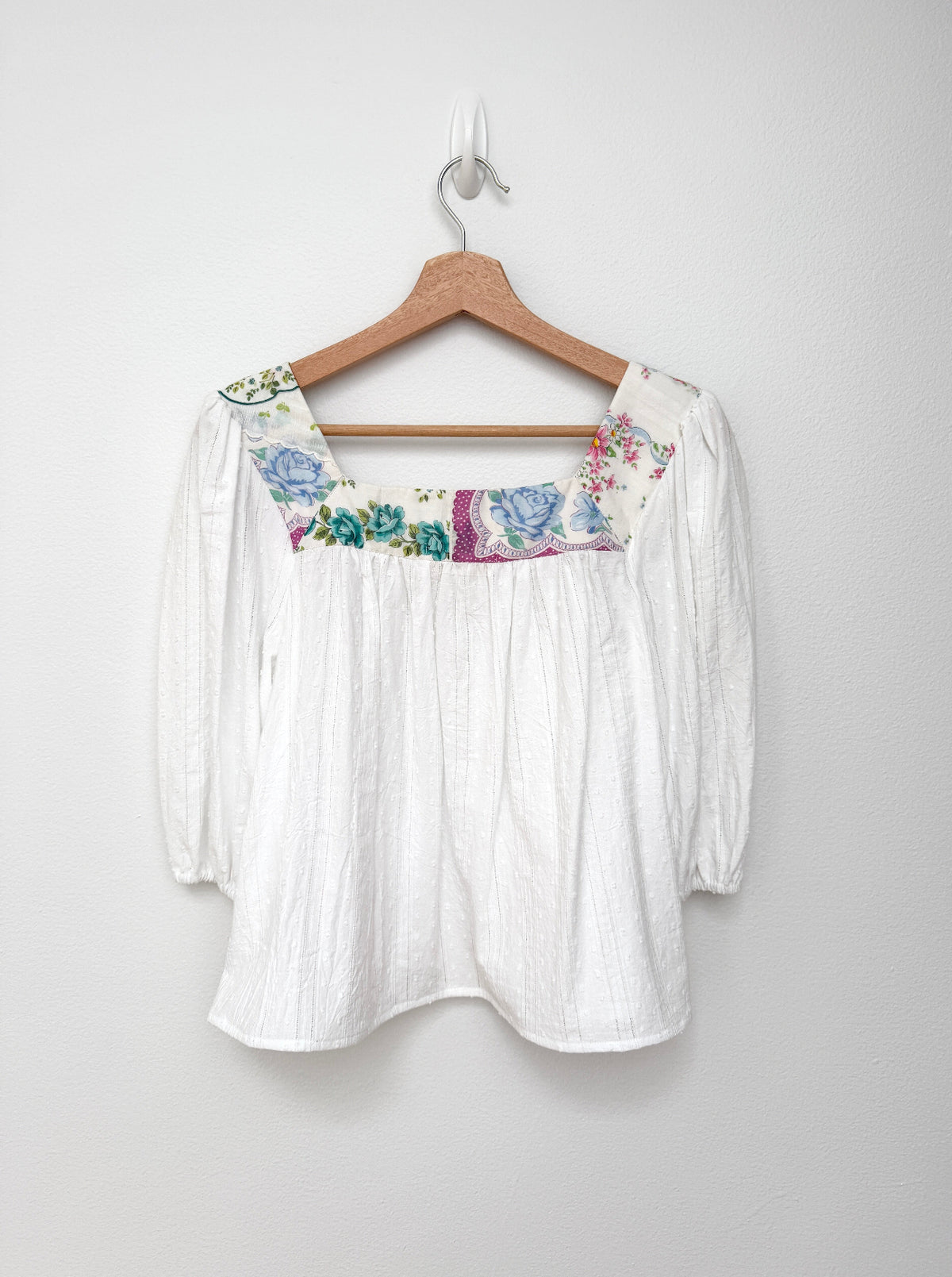 A white crinkle-textured blouse with a colorful floral embroidered yoke, hanging on a wooden hanger against a plain light gray wall.