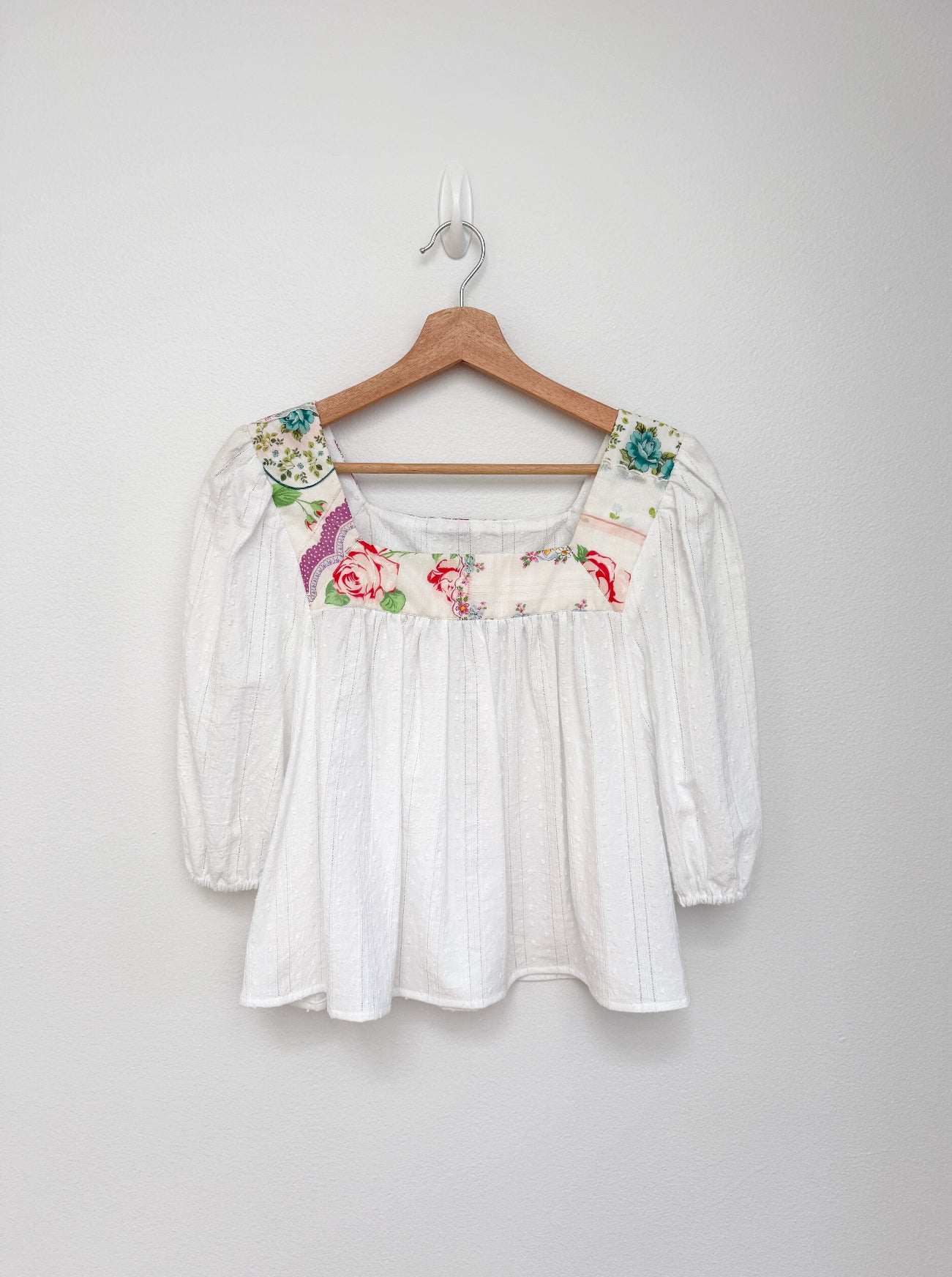 A white textured blouse with floral embroidery on the neckline and shoulders, hanging on a wooden hanger against a plain light gray wall.