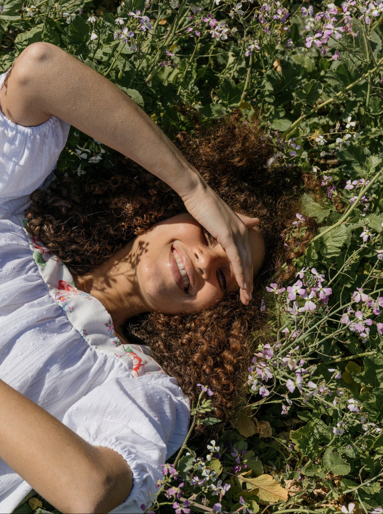 A woman lying in a field of green plants and small purple flowers, wearing a white short-sleeve blouse with a floral print at the collar.
