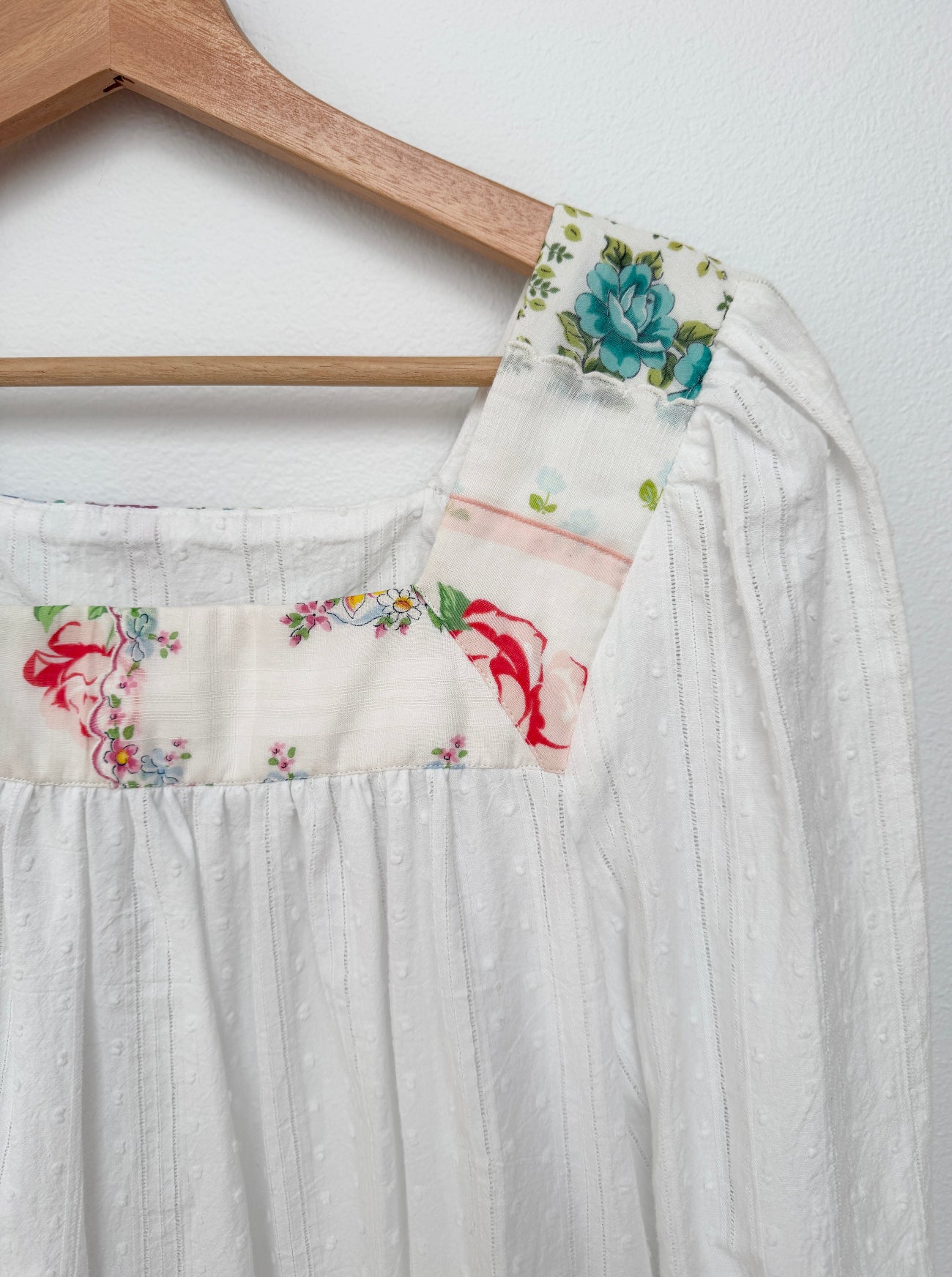 A white textured cotton dress hanging on a wooden hanger against a white wall, featuring decorative floral printed trim panels on the shoulders with blue, red, and green details.