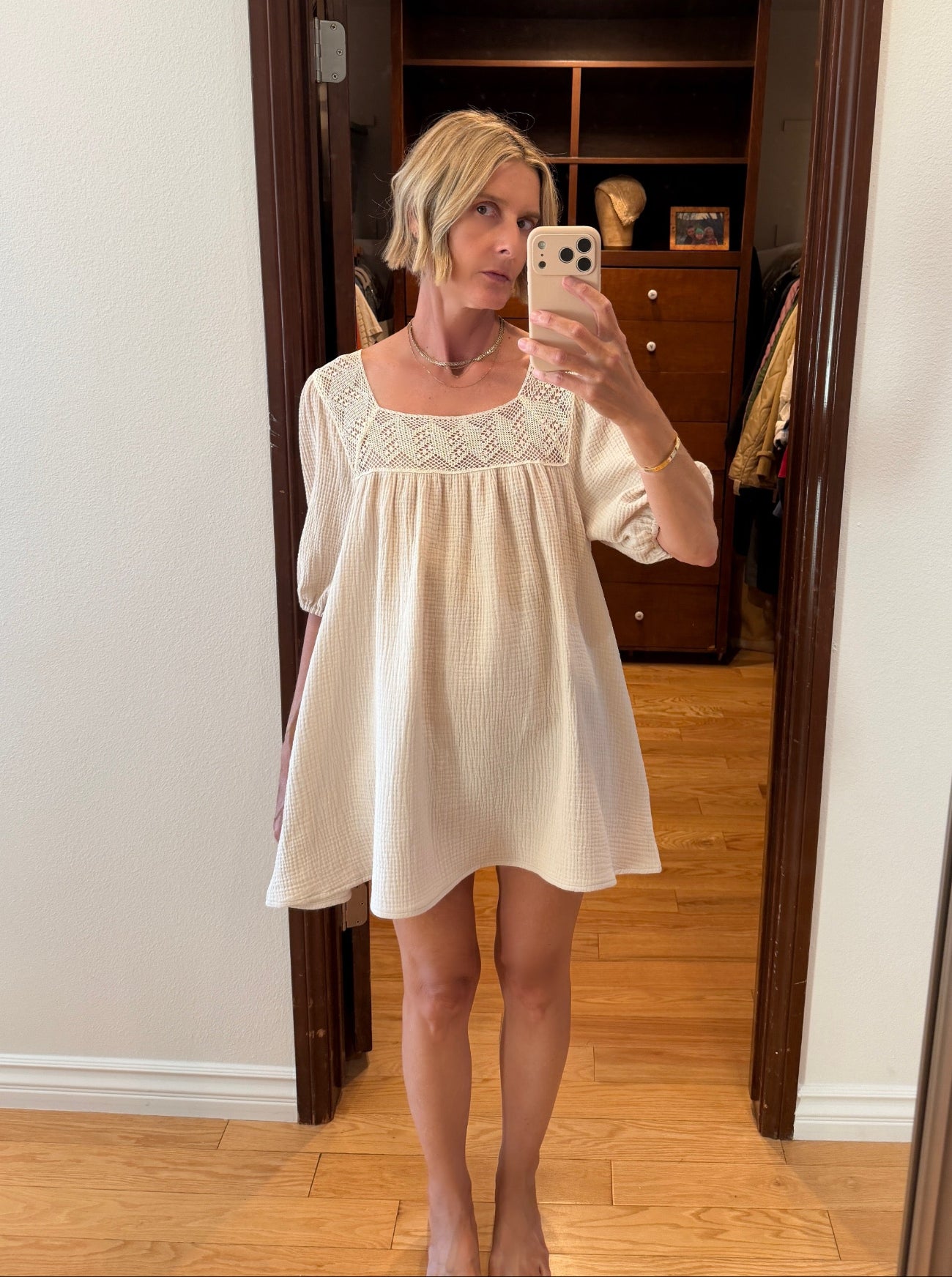 A woman takes a mirror selfie wearing a short cream-colored nightgown with a lace-trimmed square neckline. The photo is taken in a home setting with a wooden floor and a dark wood doorway behind her.