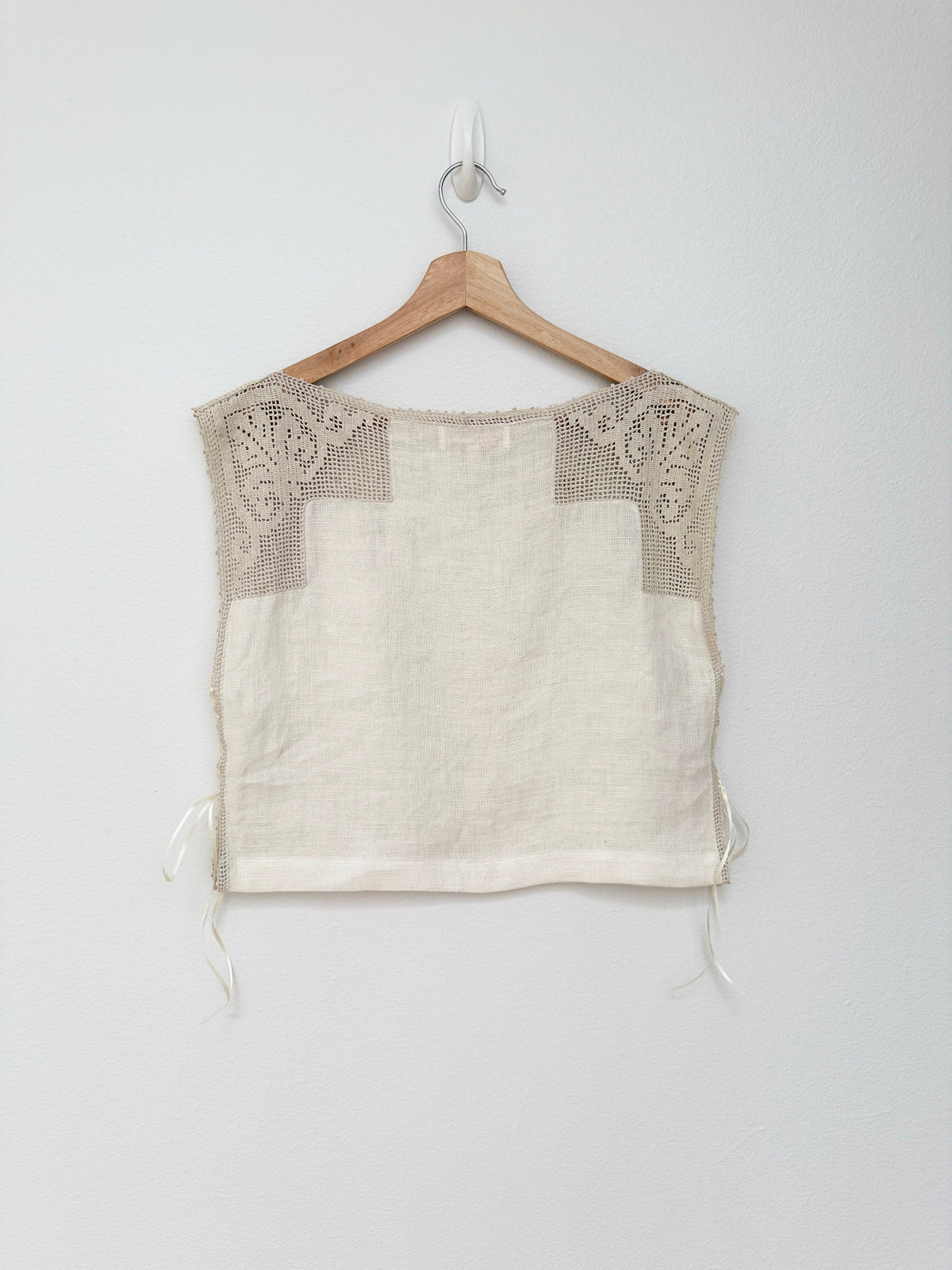 A sleeveless cropped blouse in off-white linen with beige crochet lace detailing on the shoulders, hanging on a wooden hanger against a light gray wall.