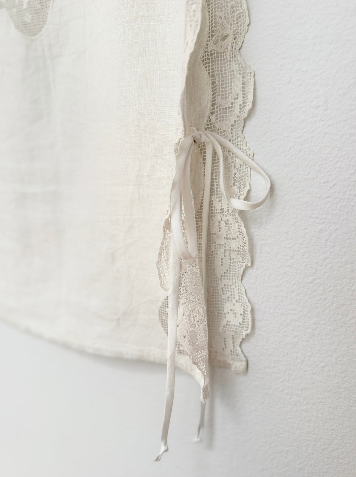 Close-up of a cream-colored textured fabric garment featuring a scalloped lace trim and matching tie details, shown against a neutral off-white background.