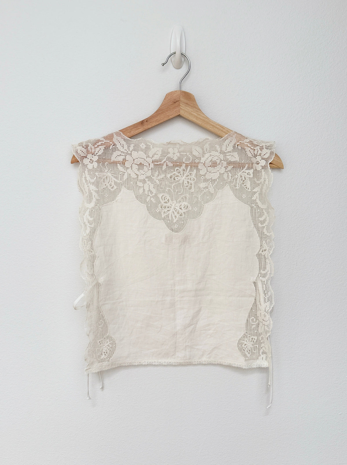 An off-white linen crop top with intricate floral lace trim, hanging on a wooden hanger against a plain light gray wall.