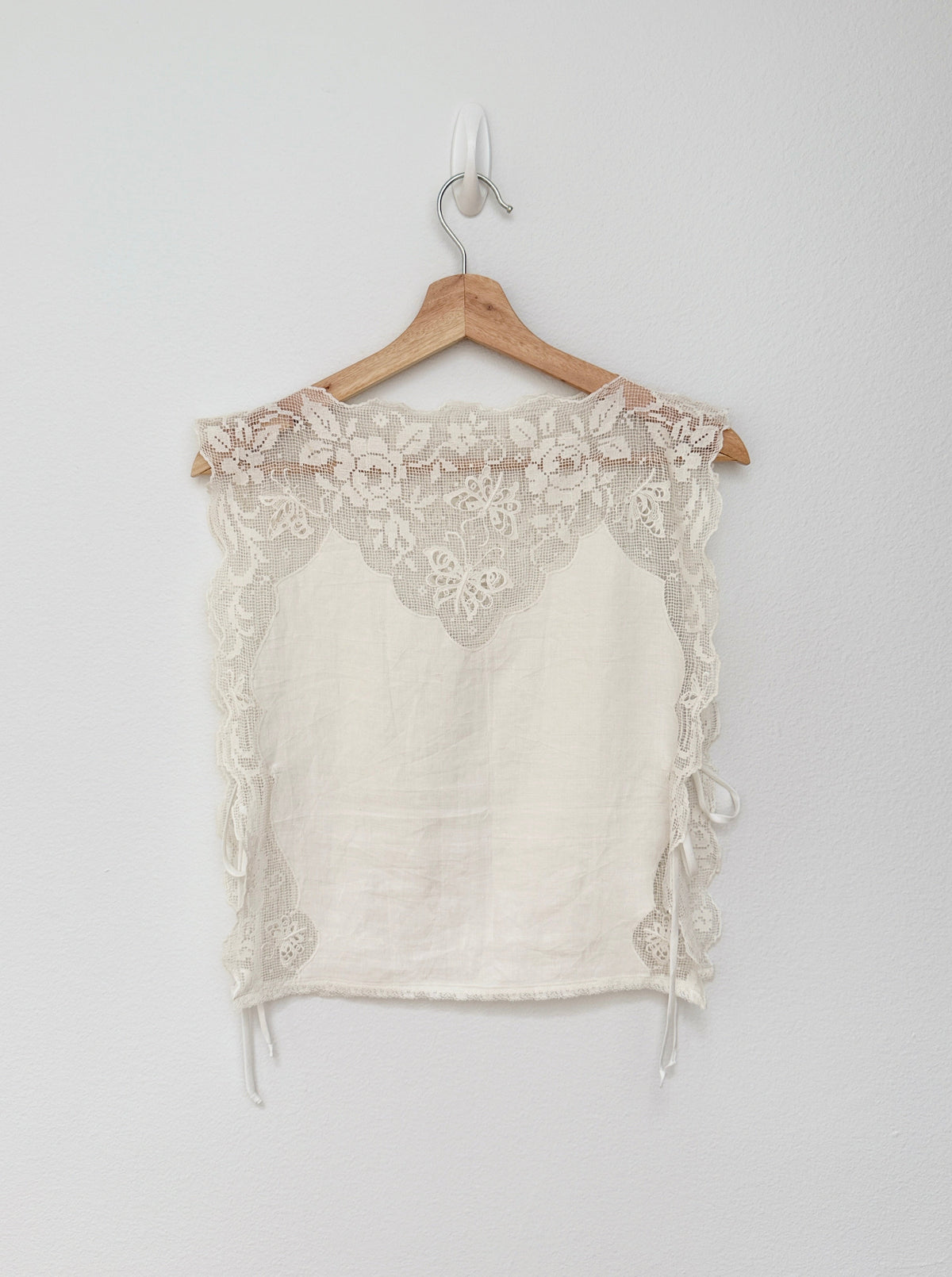 An off-white sleeveless crop top with floral lace trim and tie details, hanging on a wooden hanger against a plain light gray wall.