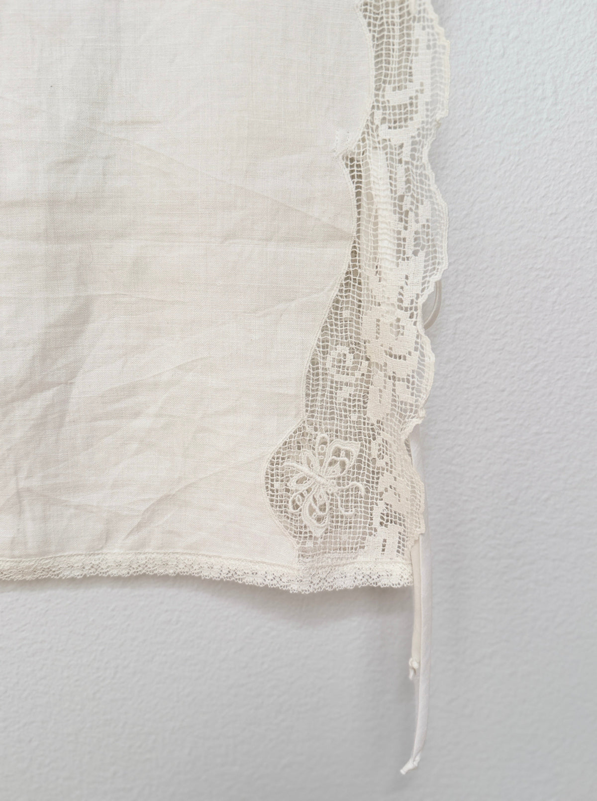 An off-white linen fabric panel with a decorative scalloped lace trim, shown hanging against a textured white wall.