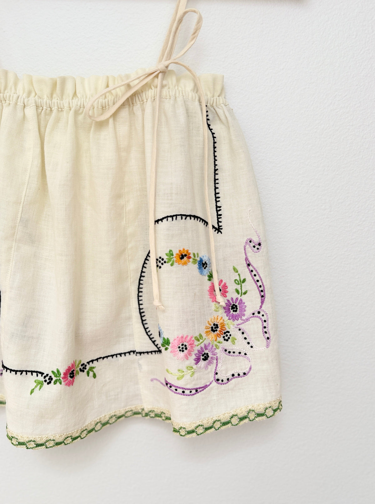 Off-white camisole top featuring colorful floral embroidery, a purple ribbon motif, and black stitched border details, hanging against a white wall.