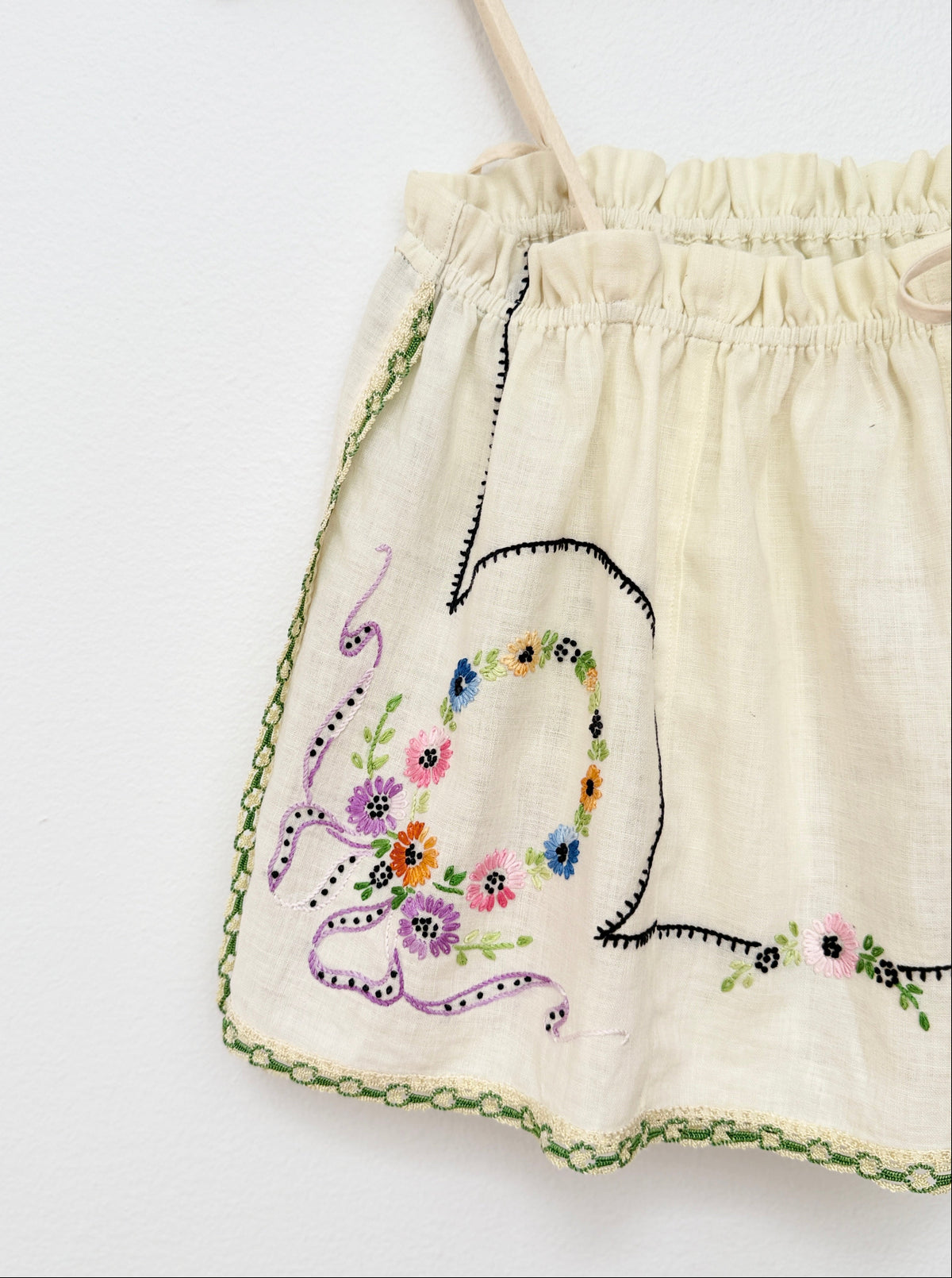 A close-up of a cream-colored embroidered top featuring a floral wreath design and green scalloped trim on a white background.