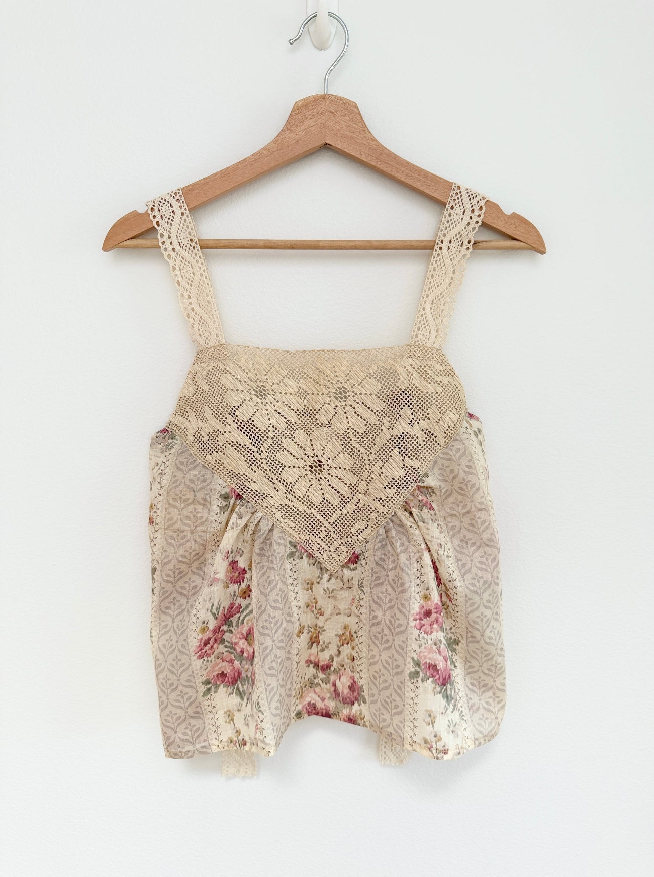 A cream lace and floral print sleeveless top hanging on a wooden hanger against a plain white wall.