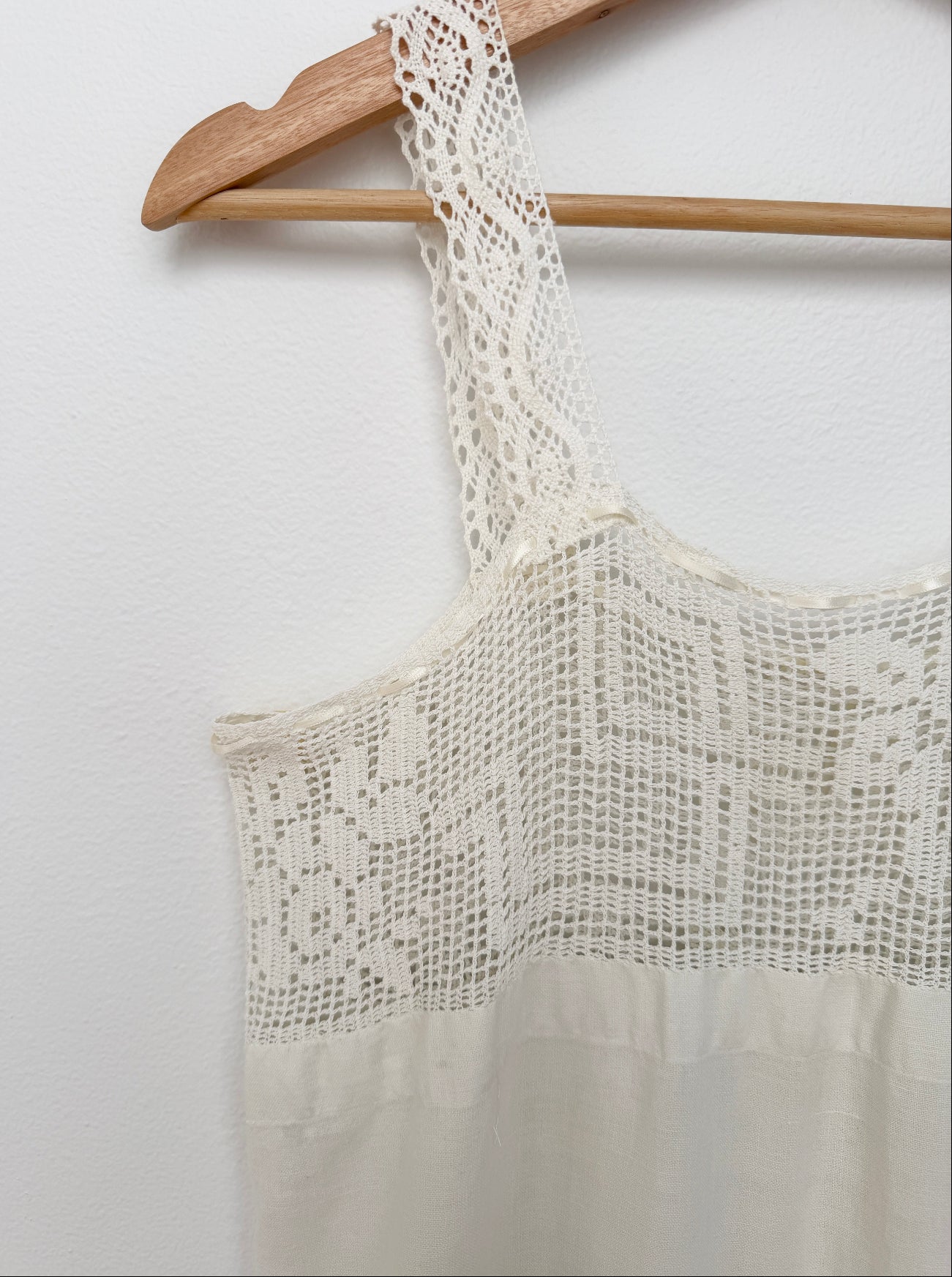 An off-white crochet knit top with a lace yoke and wide straps, hanging on a wooden hanger against a white wall.