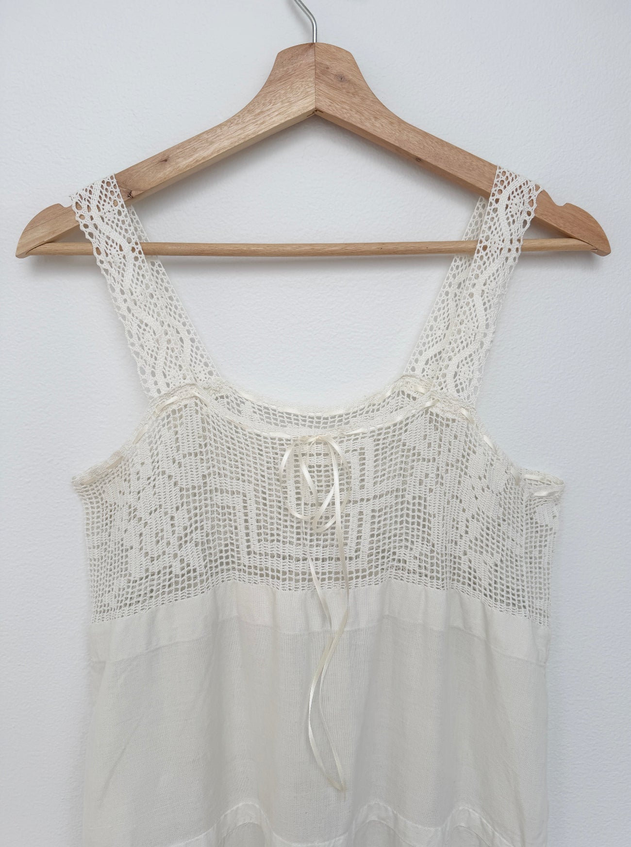 An off-white crochet and mesh camisole top hanging on a wooden hanger against a plain white wall.