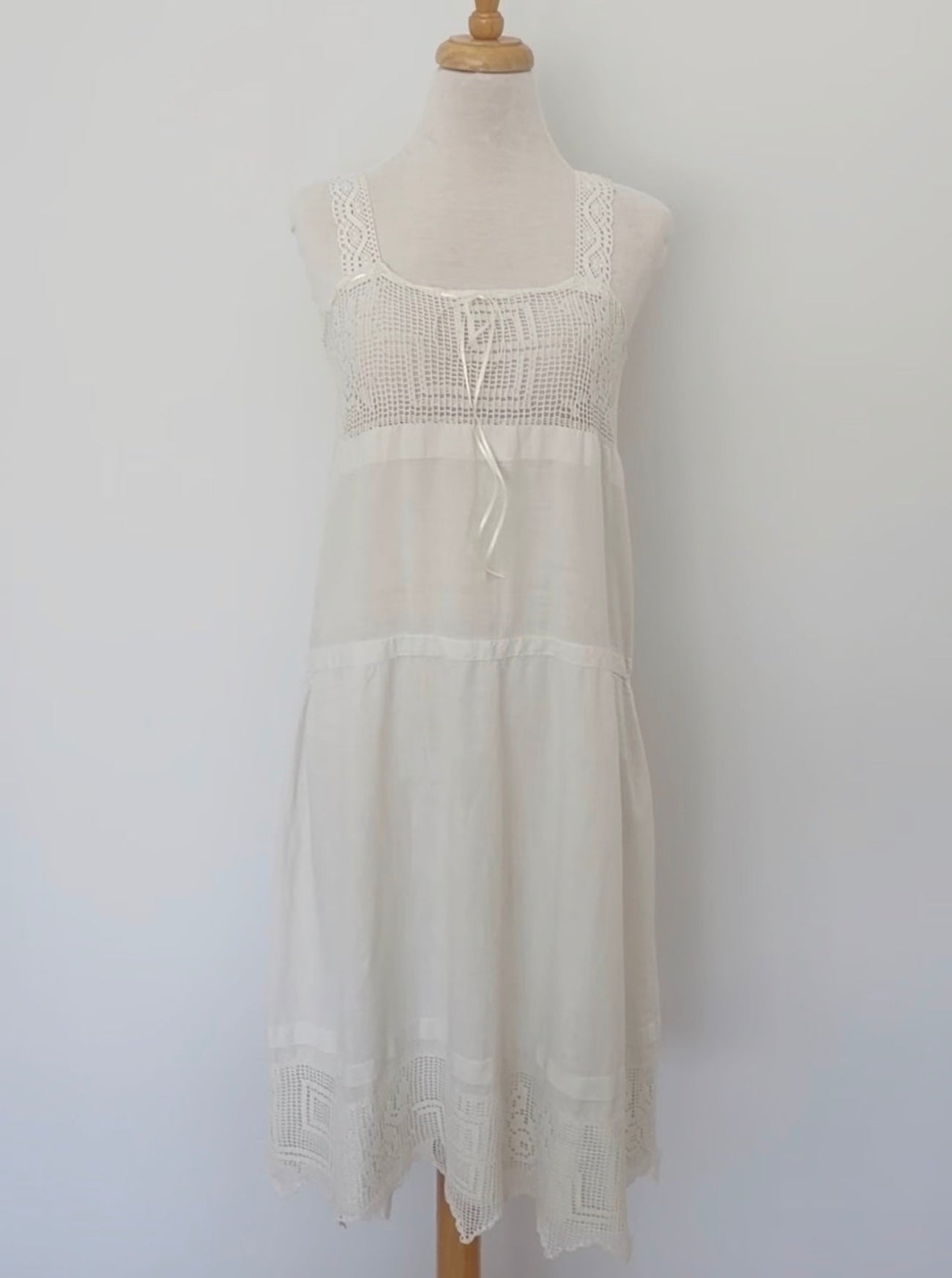 An off-white sleeveless tiered nightgown with lace trim and a front tie closure, displayed on a wooden dress form mannequin against a plain white wall.