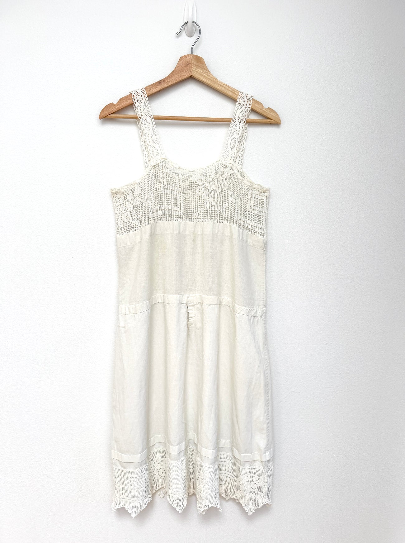 An off-white lace and embroidered sleeveless dress hanging on a wooden hanger against a plain white wall.